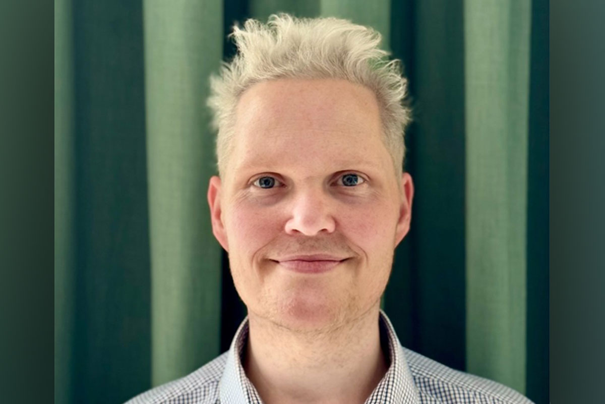 Portrait of a person with short, light blond hair and blue eyes, wearing a light checkered button-up shirt, standing in front of a green curtain and smiling gently at the camera.