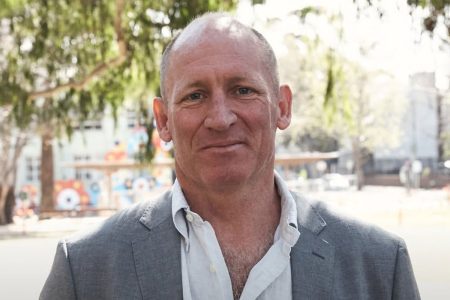 A middle-aged man stands outdoors in daylight, wearing a light grey blazer over an open-collar white shirt. He faces the camera with a neutral, slight smile, with trees and an urban streetscape softly blurred in the background.