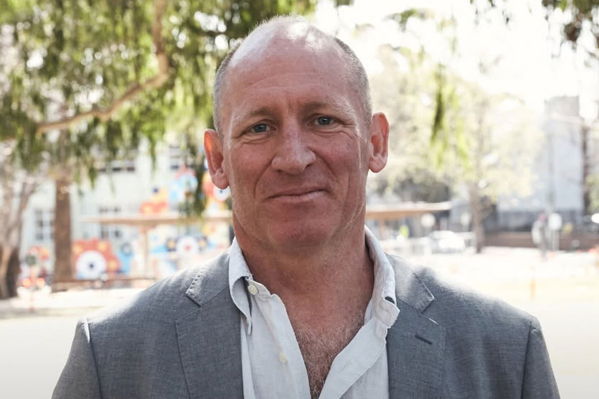 Bobby Goldsmith Foundation CEO steps down after nine years A middle-aged man stands outdoors in daylight, wearing a light grey blazer over an open-collar white shirt. He faces the camera with a neutral, slight smile, with trees and an urban streetscape softly blurred in the background.